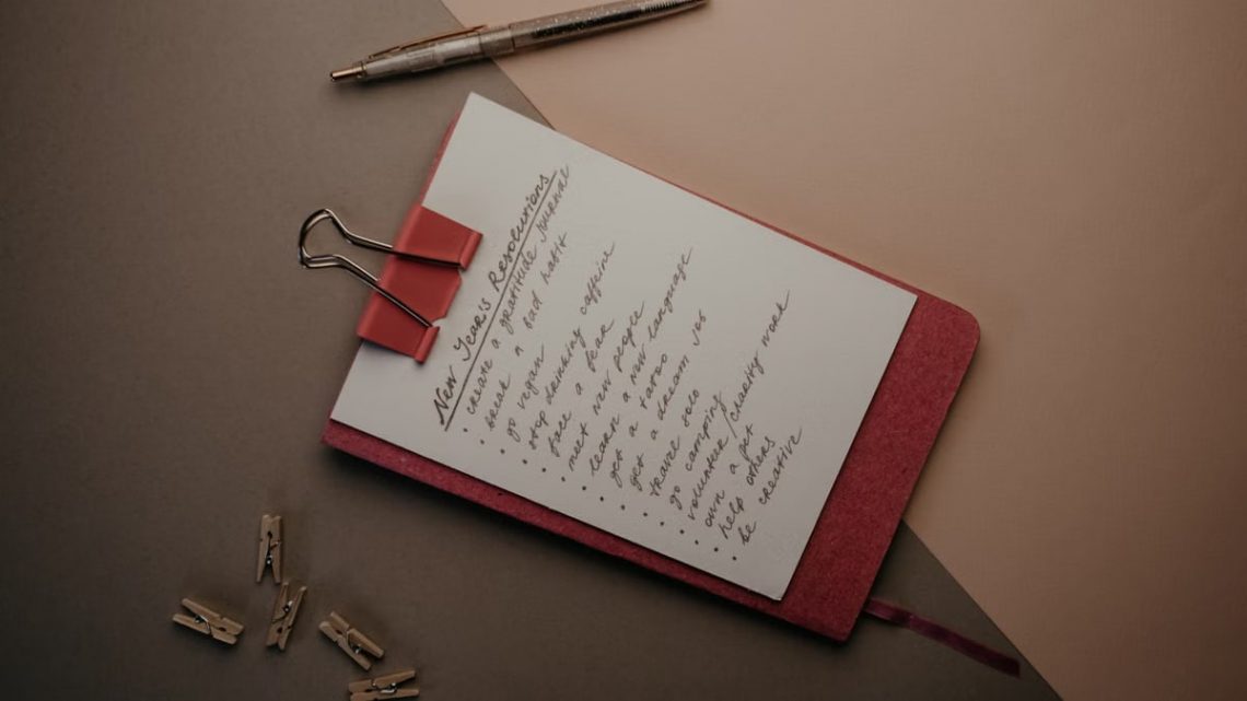 Top view of notebook with handwritten New Year's resolutions, pen, and clips on a desk.